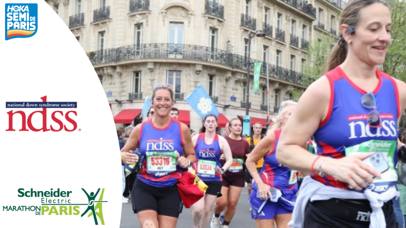 Women wearing red and blue NDSS tanks run through the streets of Paris during a marathon, smiling as they pass Parisian buildings with red awnings. Logos for HOKA Semi de Paris, National Down Syndrome Society, and Schneider Electric Marathon de Paris appear on the left.