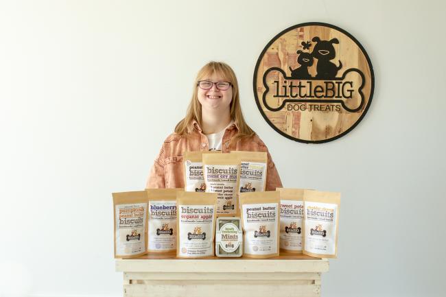 women with Down syndrome poses in front of her dog treat business logo with a table of dog treats in front her