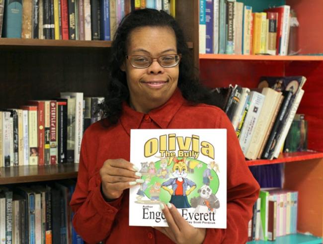 woman with down syndrome wearing black glasses and an orange long sleeve shirt holds up the book she wrote